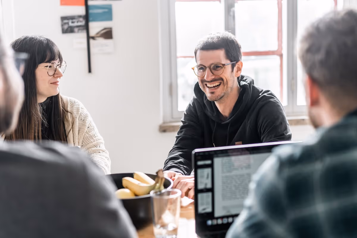 A group of young adults having a friendly conversation around a table with a laptop and bowl of bananas in a bright room.